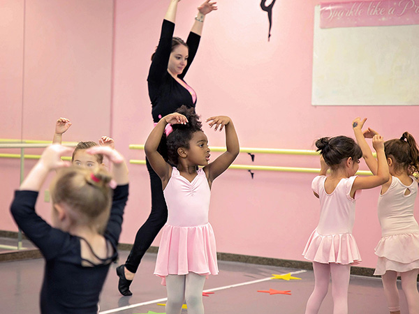 dance teacher showing little girls of various ethnicities in leotards and tutus how to do 5th position